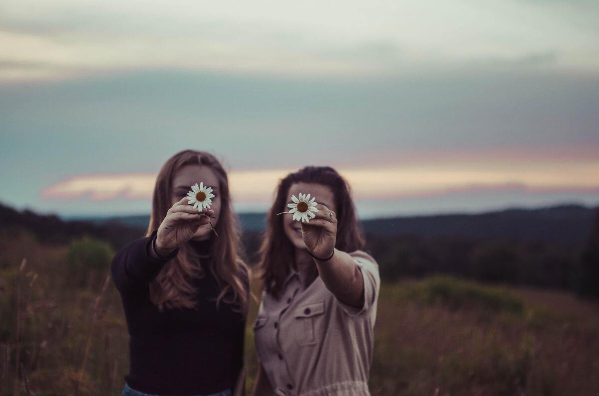 Duas mulheres segurando flores na frente de seu rosto, em um campo gramado. 