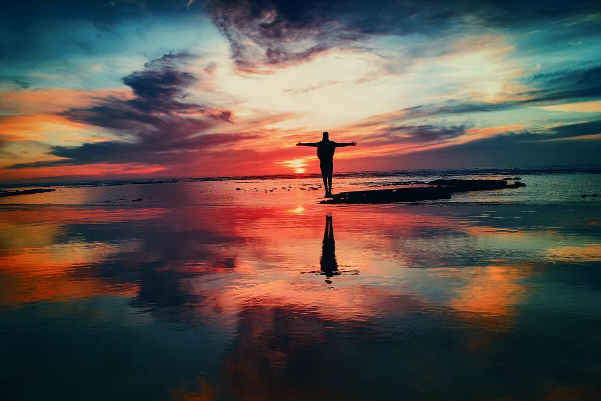 Homem de braços abertos em uma praia, pôr do sol ao fundo. 