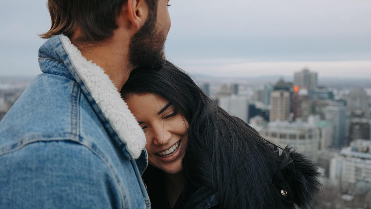 Casal abraçado, mulher sorrindo enquanto homem olha para o outro lado.