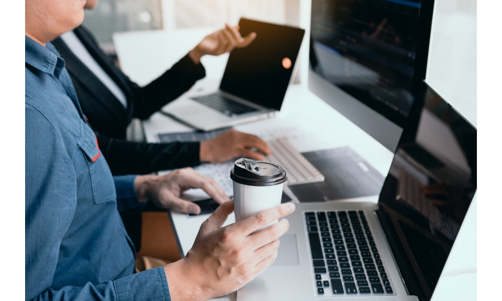 Homem trabalhando com um copo de café na mão sentado em uma mesa com notebooks e uma mulher ao fundo