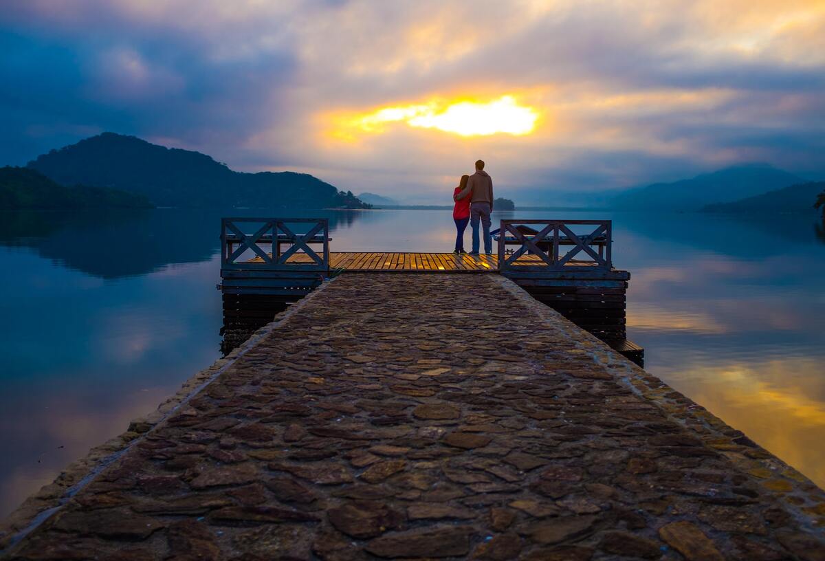 Casal abraçado em um píer, paisagem natural ao fundo.