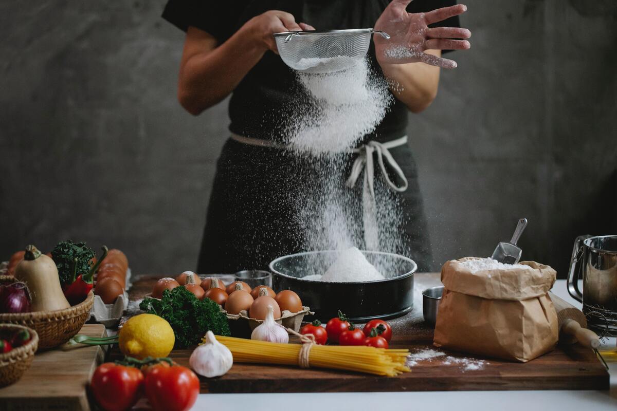 Uma mulher peneirando farinha em cima de uma mesa com muitos alimentos