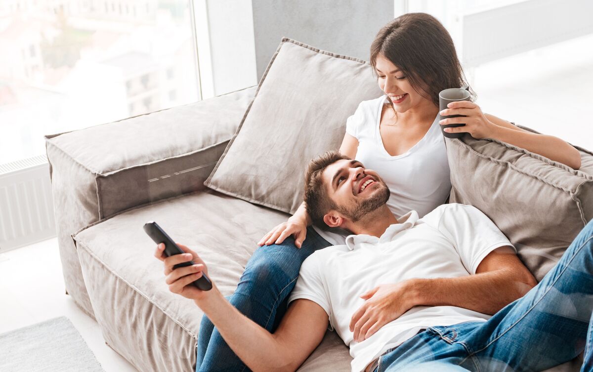 Casal de homem e mulher sentados em sofá, assistindo TV.