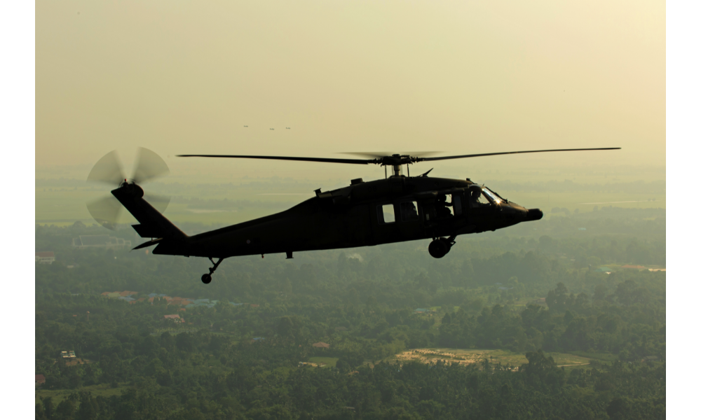 Helicóptero preto em voo com natureza ao fundo.