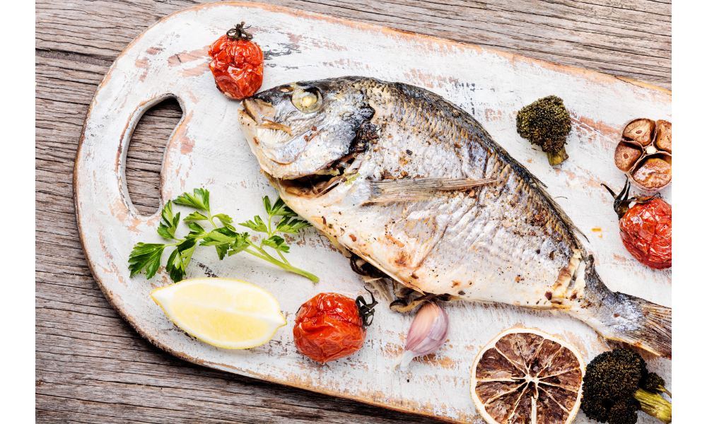 Peixe assado em uma tábua de madeira com vegetais do lado.