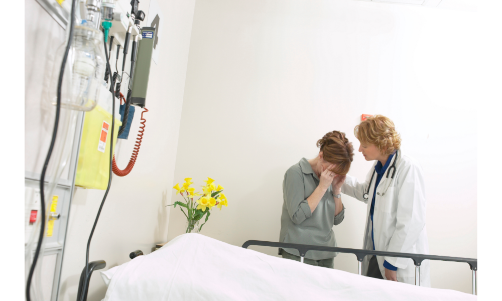 Pessoa chorando sendo consolada por uma médica atrás de uma maca vazia.