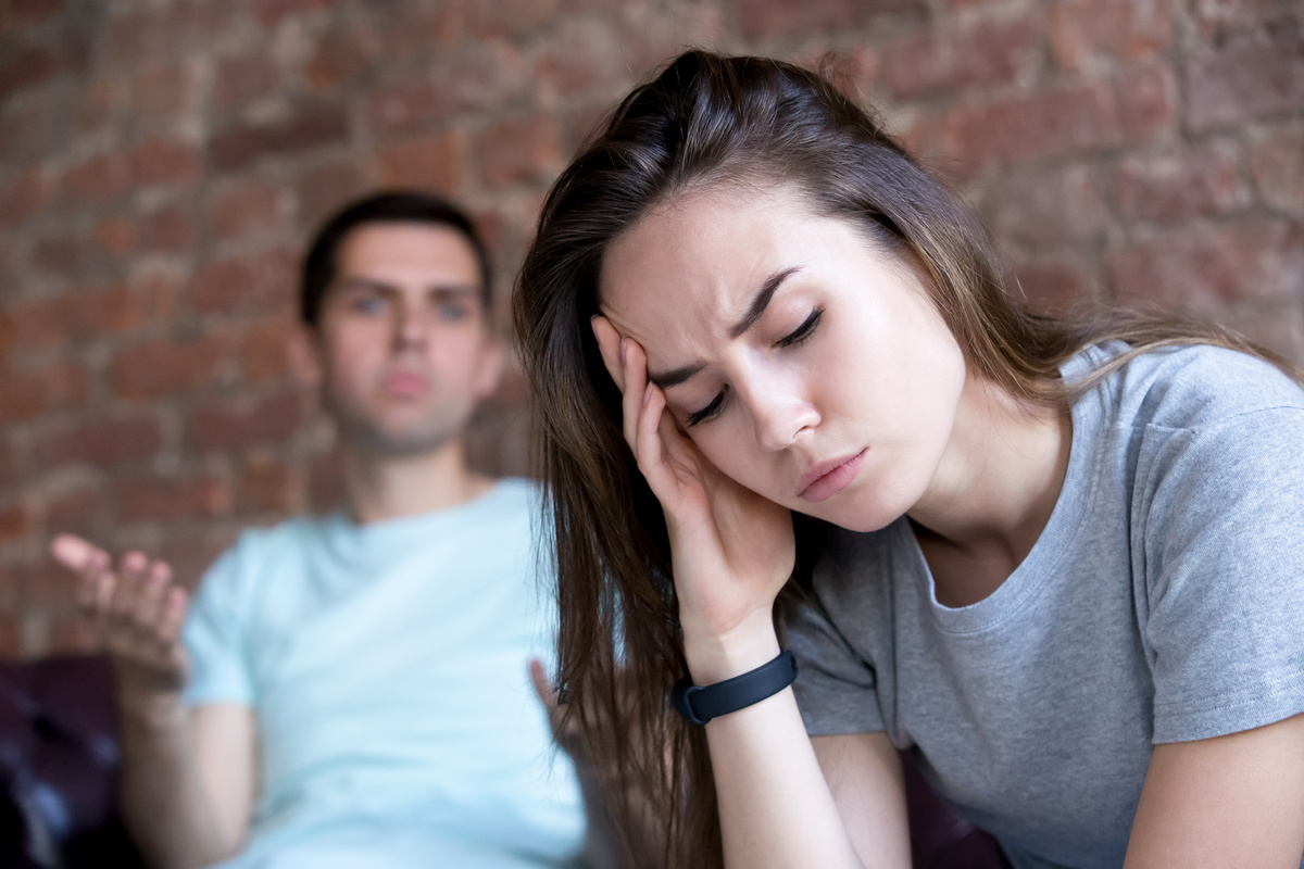 Cenário de uma briga de casal. Em primeiro plano, mulher com expressão aborrecida, de olhos fechados. Em segundo plano, logo atrás dela,homem discutindo com ela.