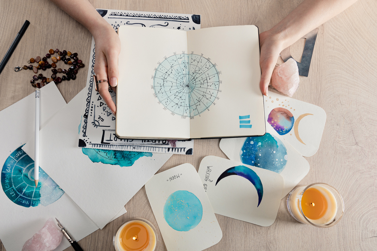 Mãos segurando um caderno com signos do zodíaco  e cartas sobre a mesa